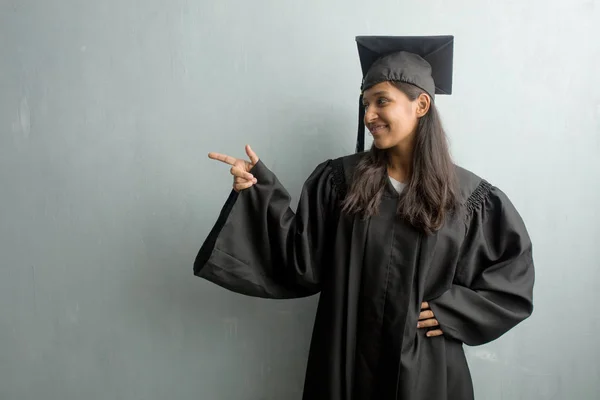 Young graduated indian woman against a wall pointing to the side, smiling surprised presenting something, natural and casual