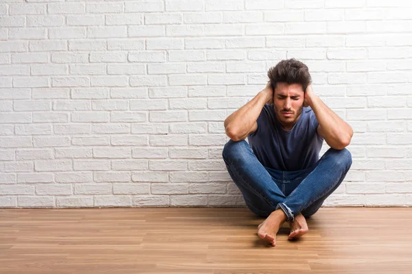 Young natural man sit on a wooden floor covering ears with hands, angry and tired of hearing some sound