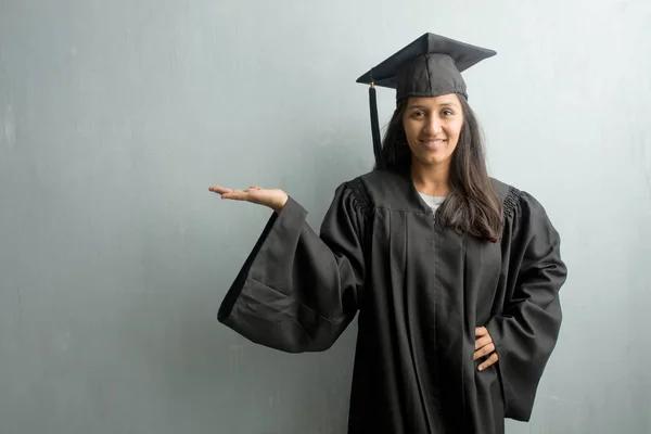 Young graduated indian woman against a wall holding something with hands, showing a product, smiling and cheerful, offering an imaginary object