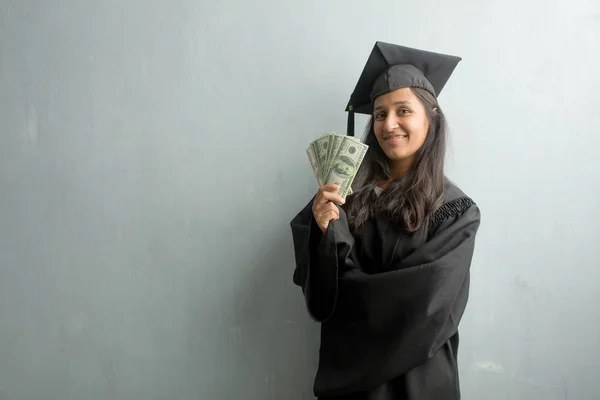 Young graduated indian woman against a wall with hands on hips, standing, relaxed and smiling, very positive and cheerful