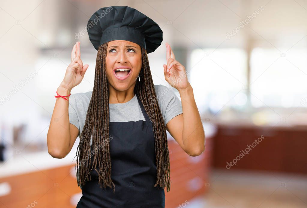 Portrait of a young black baker woman crossing his fingers, wishes to be lucky for future projects, excited but worried, nervous expression closing eyes