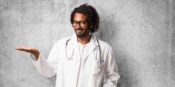 Handsome african american medical doctor holding something with hands, showing a product, smiling and cheerful, offering an imaginary object