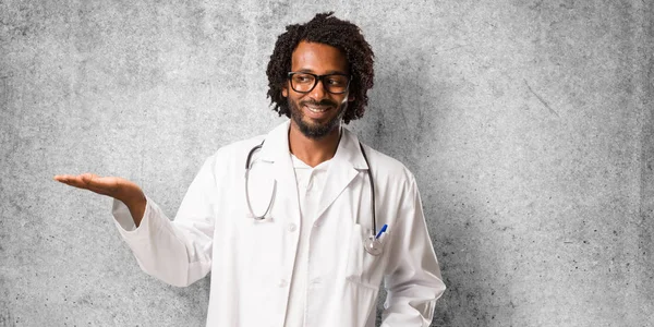 Handsome african american medical doctor holding something with hands, showing a product, smiling and cheerful, offering an imaginary object