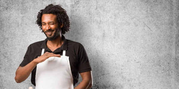 Handsome african american baker holding something with hands, showing a product, smiling and cheerful, offering an imaginary object