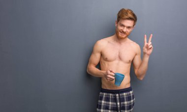 Genç gömleksiz Kızıl saçlı adam eğlenceli ve mutlu bir jest zafer yapıyor. Bir kupa kahve tutuyor.