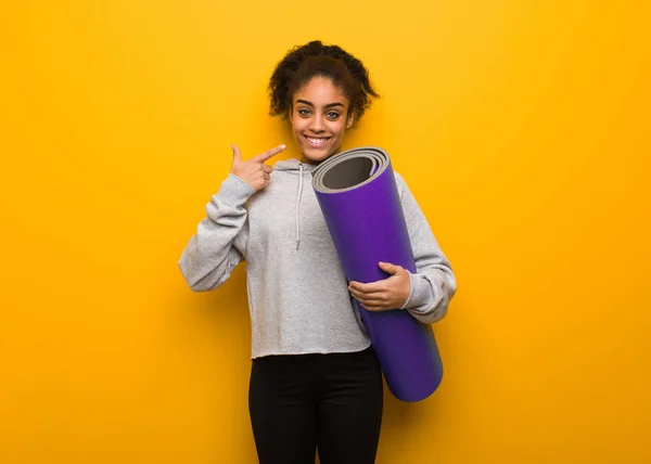 Young fitness black woman smiles, pointing mouth. Holding a mat.