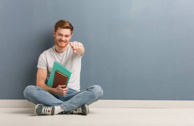 Genç Redhead öğrenci adam yerde oturan neşeli ve ön işaret gülümseyen. Kitap tutuyor..