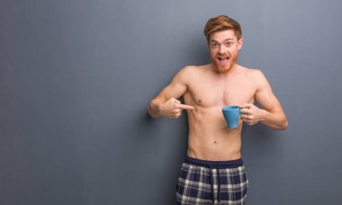 Genç gömleksiz Redhead adam şaşırttı, başarılı ve refah hissediyor. Bir kahve kupasını tutuyor..