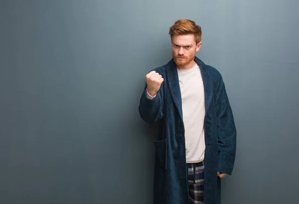 Young redhead man in pajama showing fist to front, angry expression