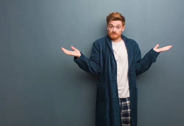 Young redhead man in pajama doubting and shrugging shoulders