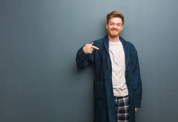 Young redhead man in pajama person pointing by hand to a shirt copy space, proud and confident