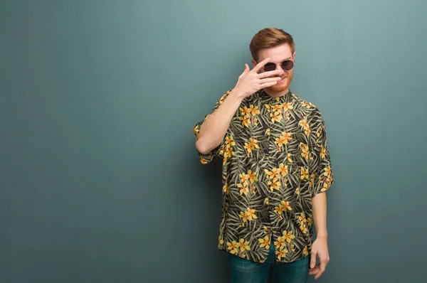 Young redhead man wearing exotic summer clothes embarrassed and laughing at the same time