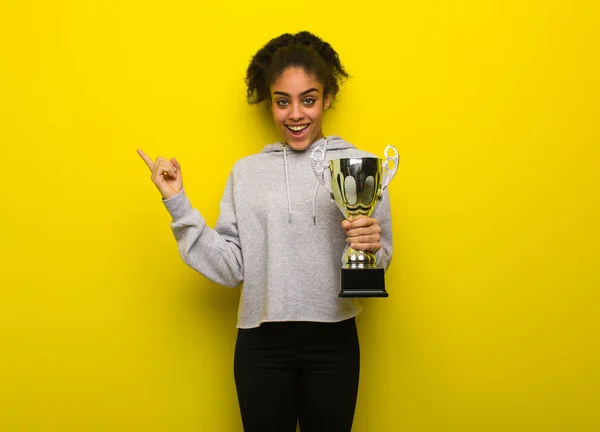 Young fitness black woman pointing to the side with finger. Holding a trophy.