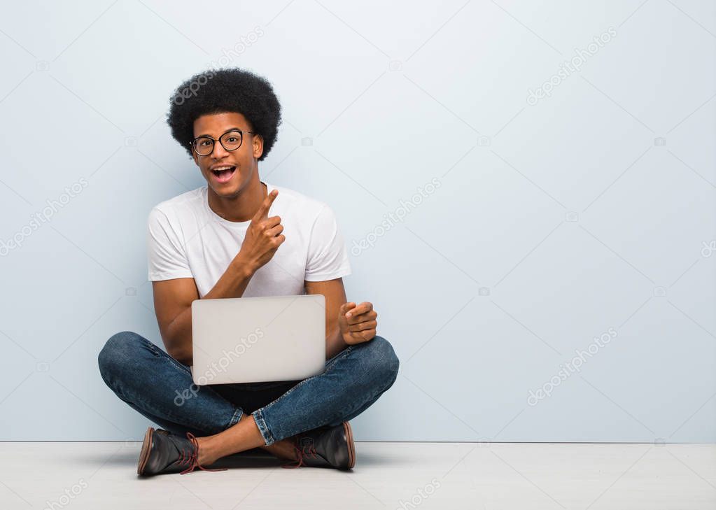 Young black man sitting on the floor with a laptop pointing to the side with finger