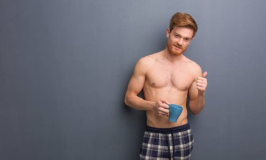 Young shirtless redhead man smiling and raising thumb up. He is holding a coffee mug. clipart