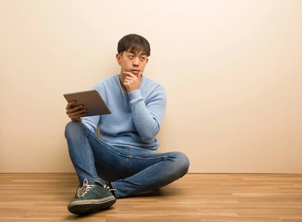 Young chinese man sitting using his tablet doubting and confused
