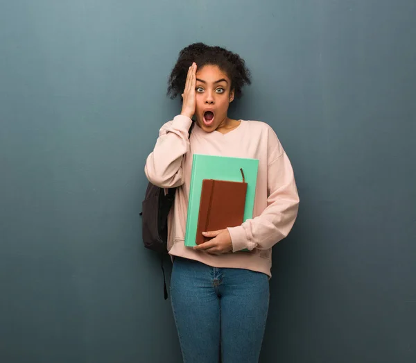 Young student black woman surprised and shocked. She is holding books.