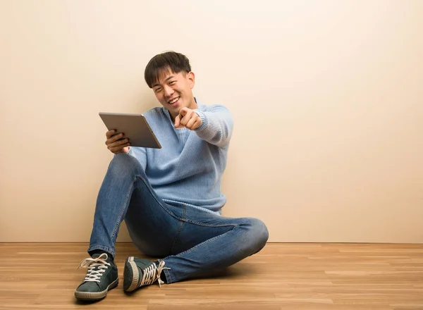 年轻的中国男子坐在用他的平板电脑欢快和微笑指向前面 — 图库照片