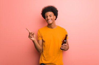 Genç Afro-Amerikalı bir adam elinde bira tutarken parmağıyla kenarı işaret ediyor.