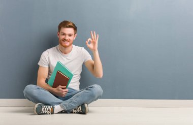 Genç kızıl saçlı öğrenci yerde neşeli ve kendinden emin bir şekilde oturuyor ve iyi bir jest yapıyor. Elinde kitaplar var..