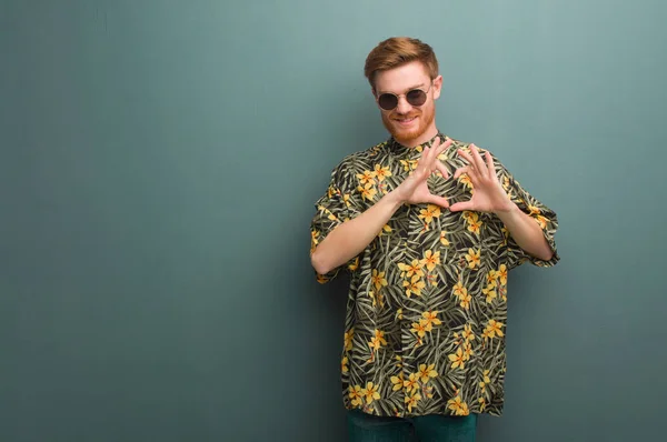 Young redhead man wearing exotic summer clothes doing a heart shape with hands
