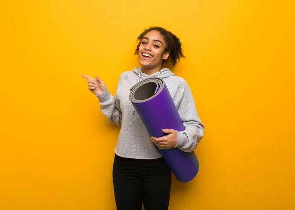 Young fitness black woman pointing to the side with finger. Holding a mat.