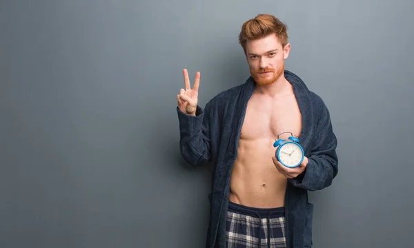 Young redhead man wearing pajama fun and happy doing a gesture of victory. He is holding an alarm clock.