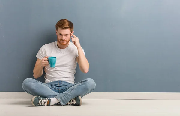 Young redhead student man sitting on the floor thinking about an idea. He is holding a coffee mug.