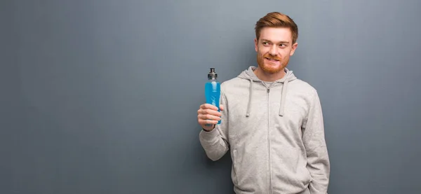 Young redhead fitness man smiling confident and crossing arms, looking up. He is holding an energy drink.
