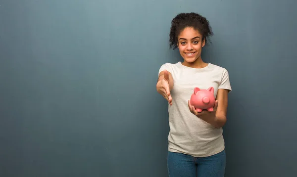 Young black woman reaching out to greet someone. She is holding a piggy bank.