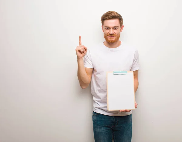 Young redhead man showing number one. He is holding a clipboard.