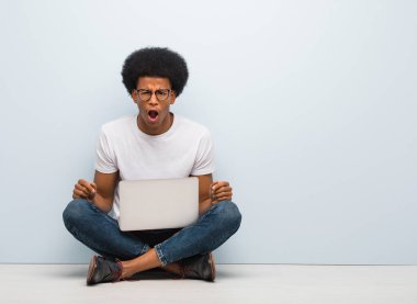 Genç siyah bir adam dizüstü bilgisayarla yerde oturuyor ve çok sinirli ve agresif bir şekilde bağırıyor.