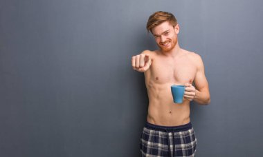 Genç, üstsüz, kızıl saçlı, neşeli ve gülümseyen adam öne bakıyor. Elinde bir kahve fincanı var..