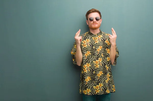 Young redhead man wearing exotic summer clothes crossing fingers for having luck