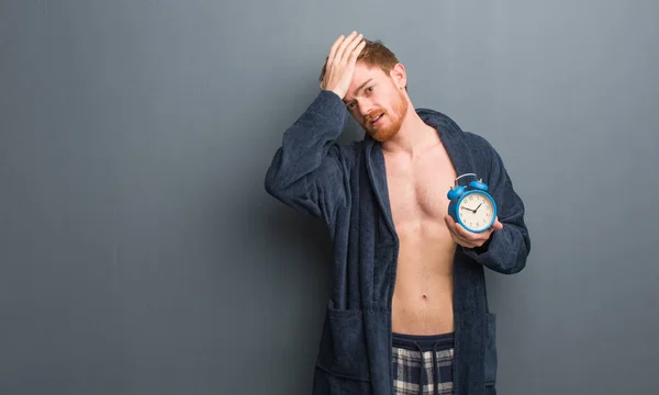 Young redhead man wearing pajama worried and overwhelmed. He is holding an alarm clock.