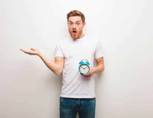 Young redhead man holding something on palm hand. He is holding an alarm clock.