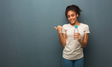 Genç siyah kadın gülümsüyor ve yan tarafı işaret ediyor. Elinde bir su şişesi var..