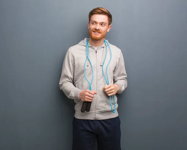 Young redhead fitness man smiling confident and crossing arms, looking up. He is holding a jump rope.