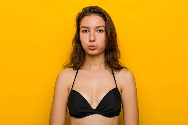 Young european woman wearing bikini blows cheeks, has tired expression. Facial expression concept.