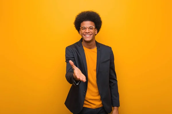 Young business african american man over an orange wall reaching out to greet someone