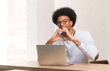 Genç siyah adam dizüstü bilgisayarını kullanıyor. Elleriyle kalp şekli yapıyor.