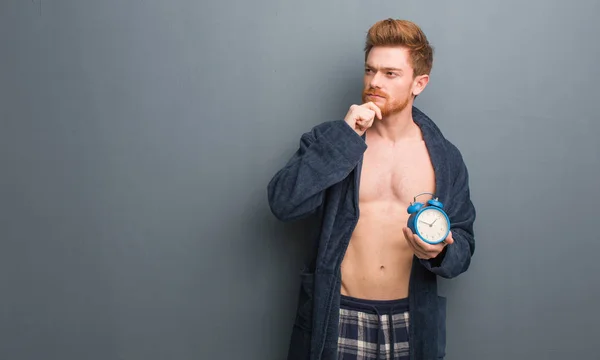 Young redhead man wearing pajama doubting and confused. He is holding an alarm clock.
