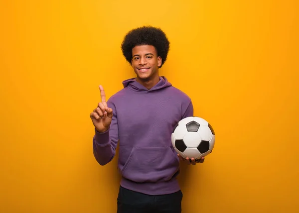 Young african american sport man holding a soccer ball showing number one