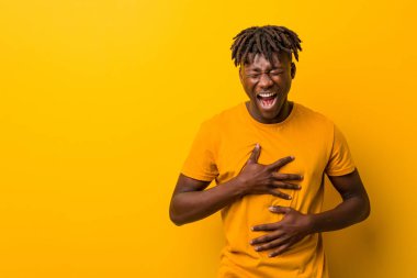 Sarı arka planda rastalar takan genç siyahi adam mutlu bir şekilde gülüyor ve ellerini karnının üstünde tutarak eğleniyor..