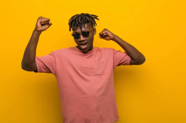 Young casual black man wearing sunglasses celebrating a special day, jumps and raising arms with energy.