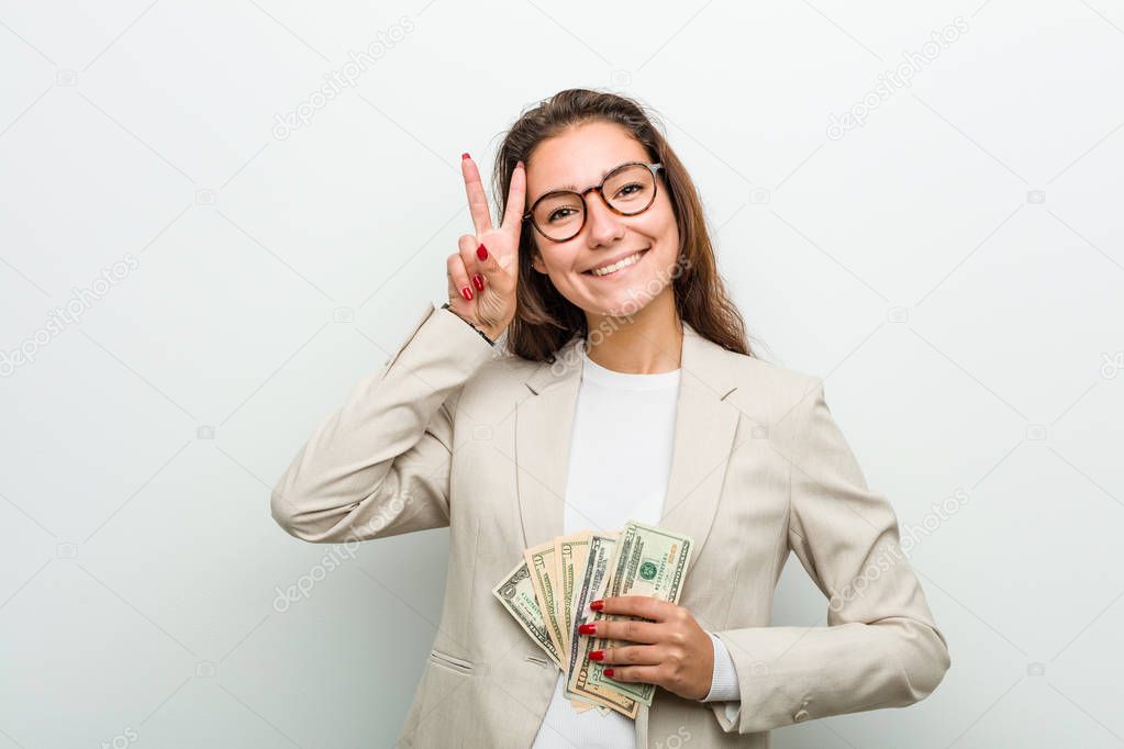 Young european business woman holding dollar banknotes showing victory sign and smiling broadly.