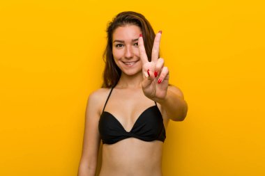 Bikini giyen genç Avrupalı kadın iki numarayı parmaklarıyla gösteriyor..