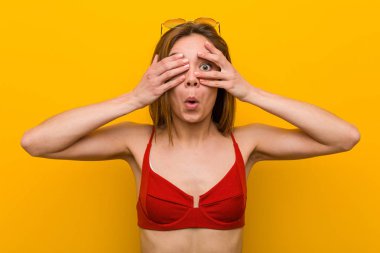 Bikini giyen ve güneş gözlüğü takan beyaz bir kadın parmaklarının arasından korkmuş ve gergin bir şekilde göz kırpıyor..
