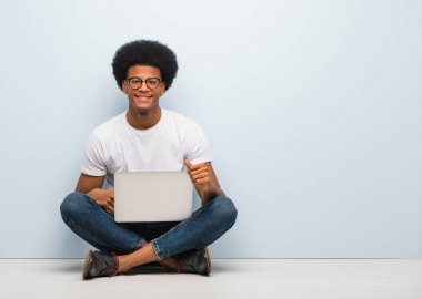 Genç siyahi bir adam dizüstü bilgisayarıyla yerde oturuyor. Kocaman bir gülümsemesi var.