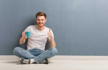 Genç kızıl saçlı öğrenci yerde oturmuş gülümsüyor ve başparmağını kaldırıyor. Elinde bir kahve fincanı var..